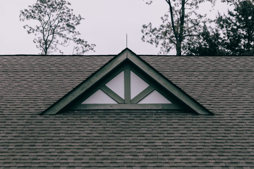 A triangular attic vent with green trim is centered near the roof peak of a shingled building, with tall trees and an overcast sky framing the roofline in the background.