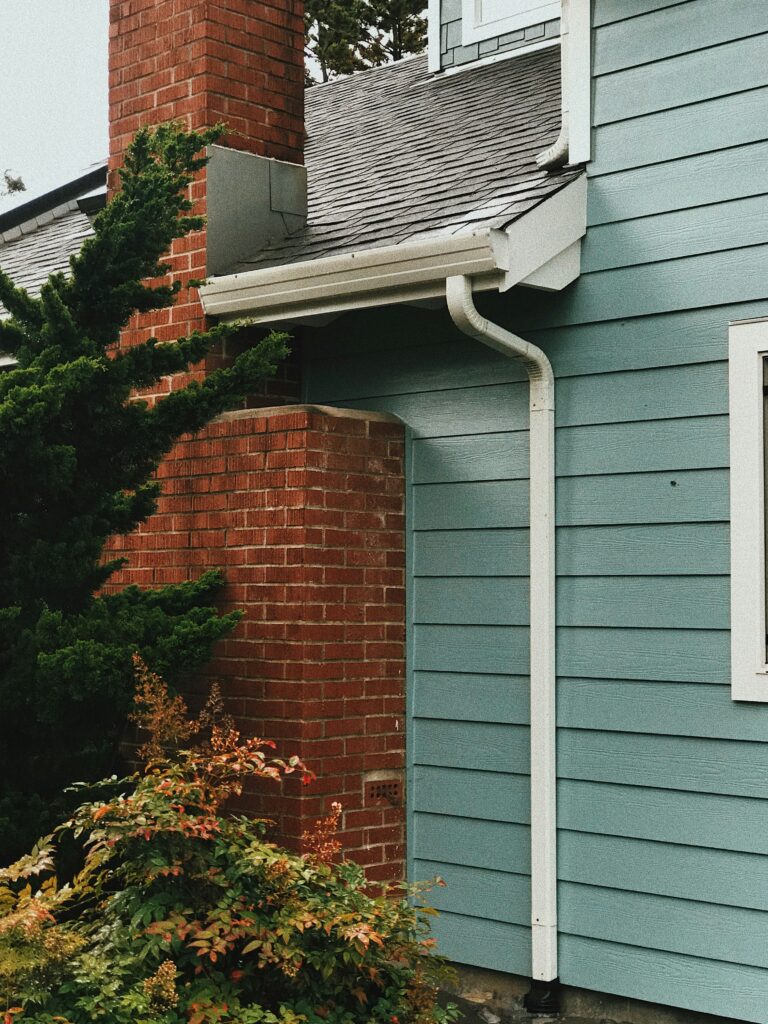 A close-up view of a house exterior shows blue siding, a brick chimney, green shrubs in the foreground, and well-maintained gutters along the white gutter system.