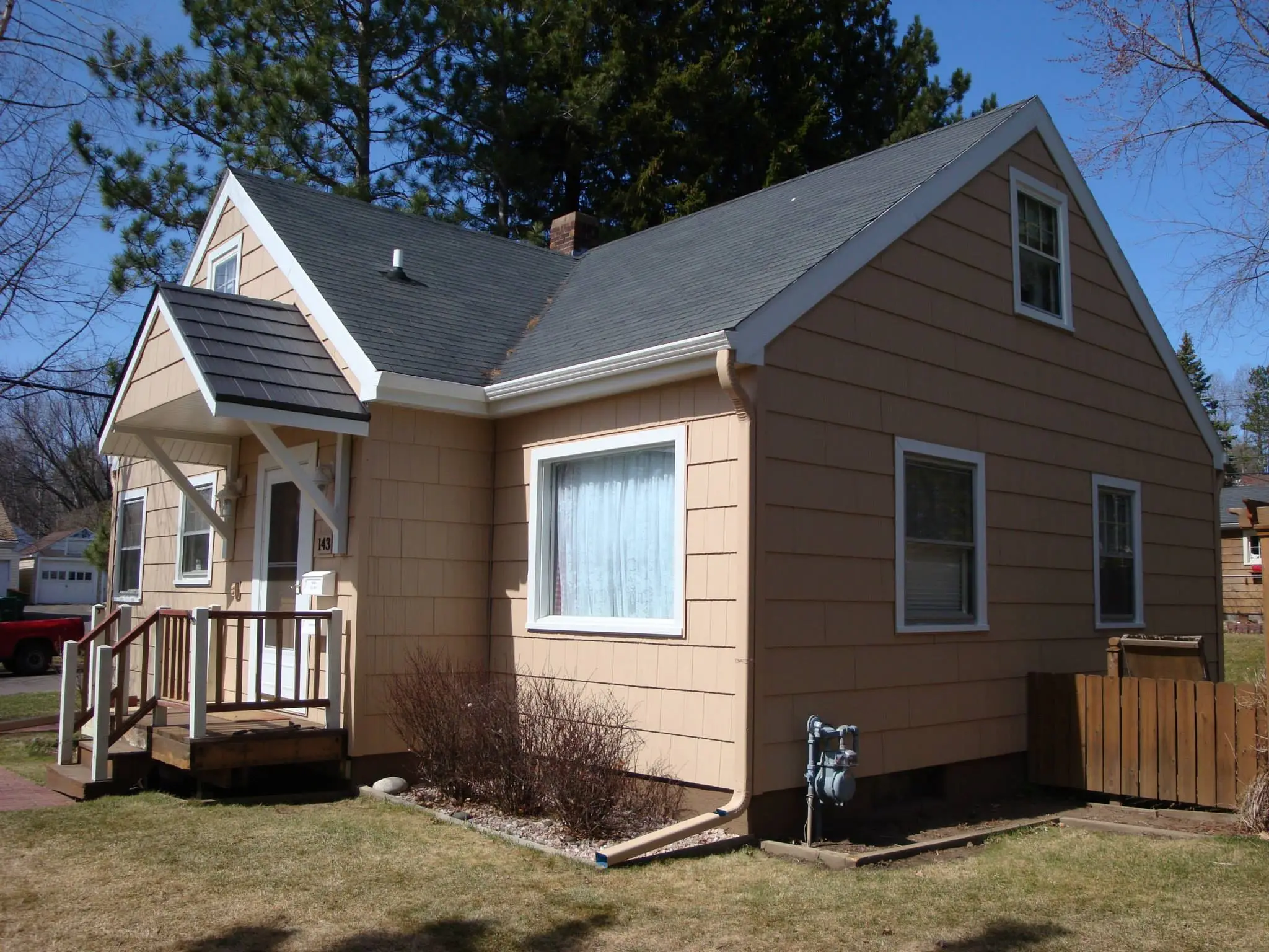 A small beige house with white trim, a covered front porch, and multiple windows sits in a yard with sparse grass and bare shrubs—an ideal candidate for exterior renovation. Tall pine trees rise in the background.
