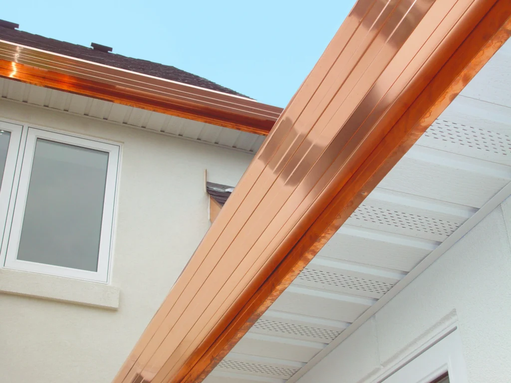 Close-up view of a house exterior featuring shiny copper gutters installed along the roof’s edge, with white soffits and light-colored walls highlighted under a clear blue sky.