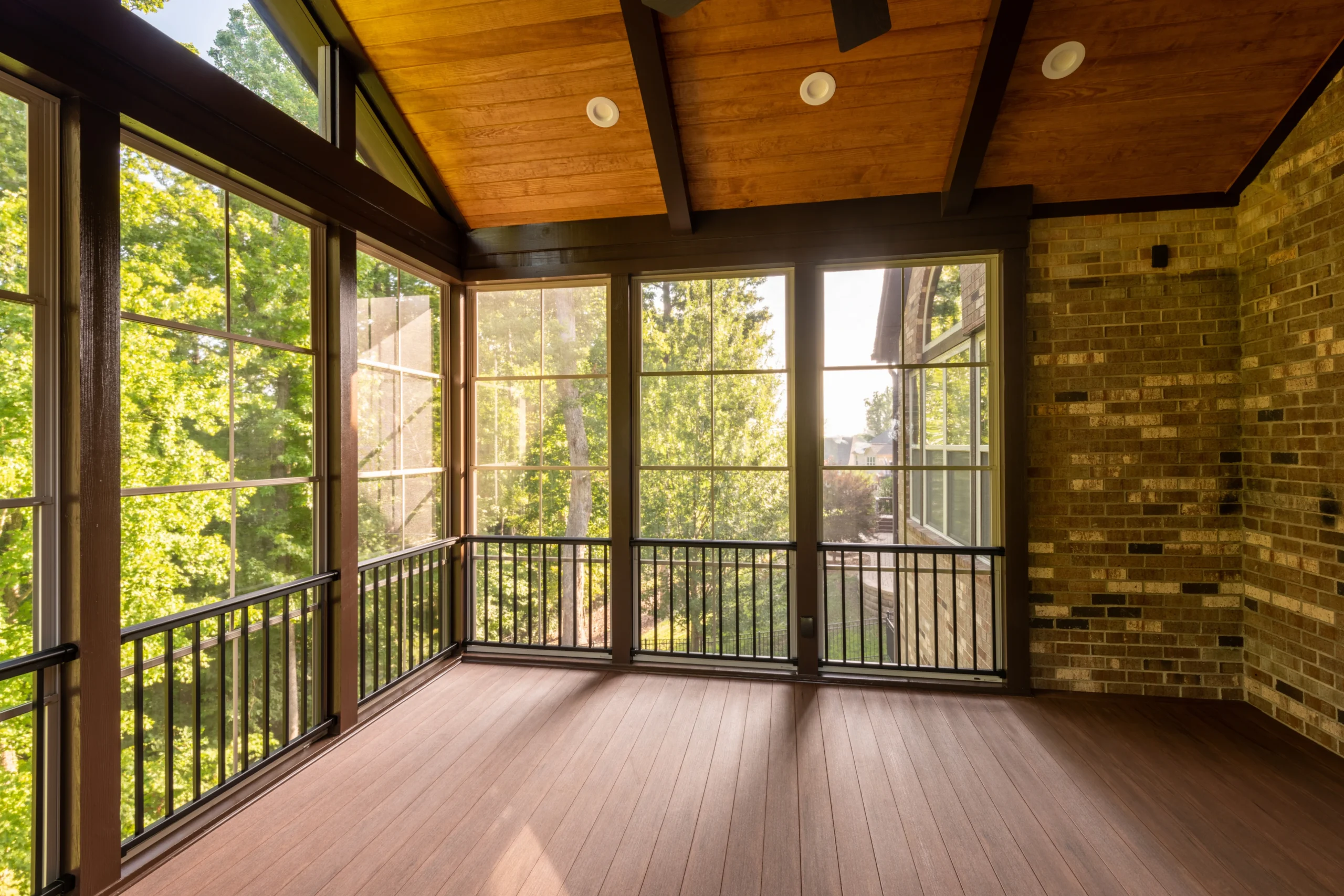 A spacious screened-in patio with wood flooring, a vaulted wooden ceiling, large windows that invite natural light, and black metal railings, overlooking green trees and brick exterior walls.