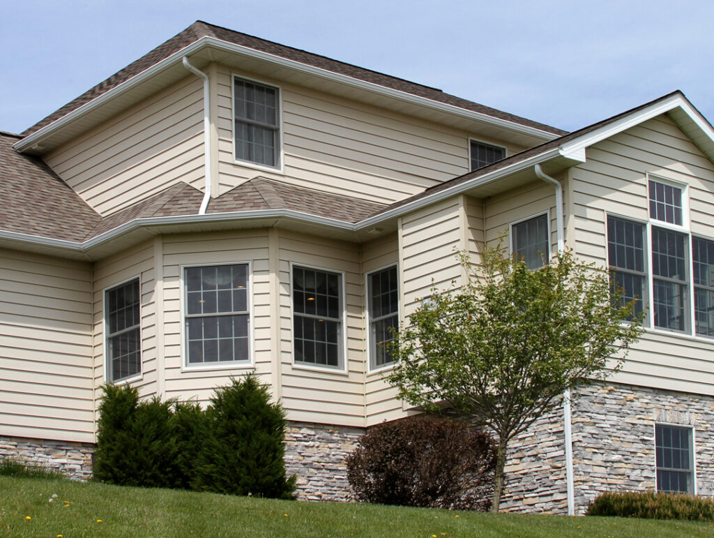 Two-story suburban house with vinyl siding and stone accents, multiple large windows, a gable roof, small bushes, and a tree in the front yard on a grassy lawn under a clear sky.