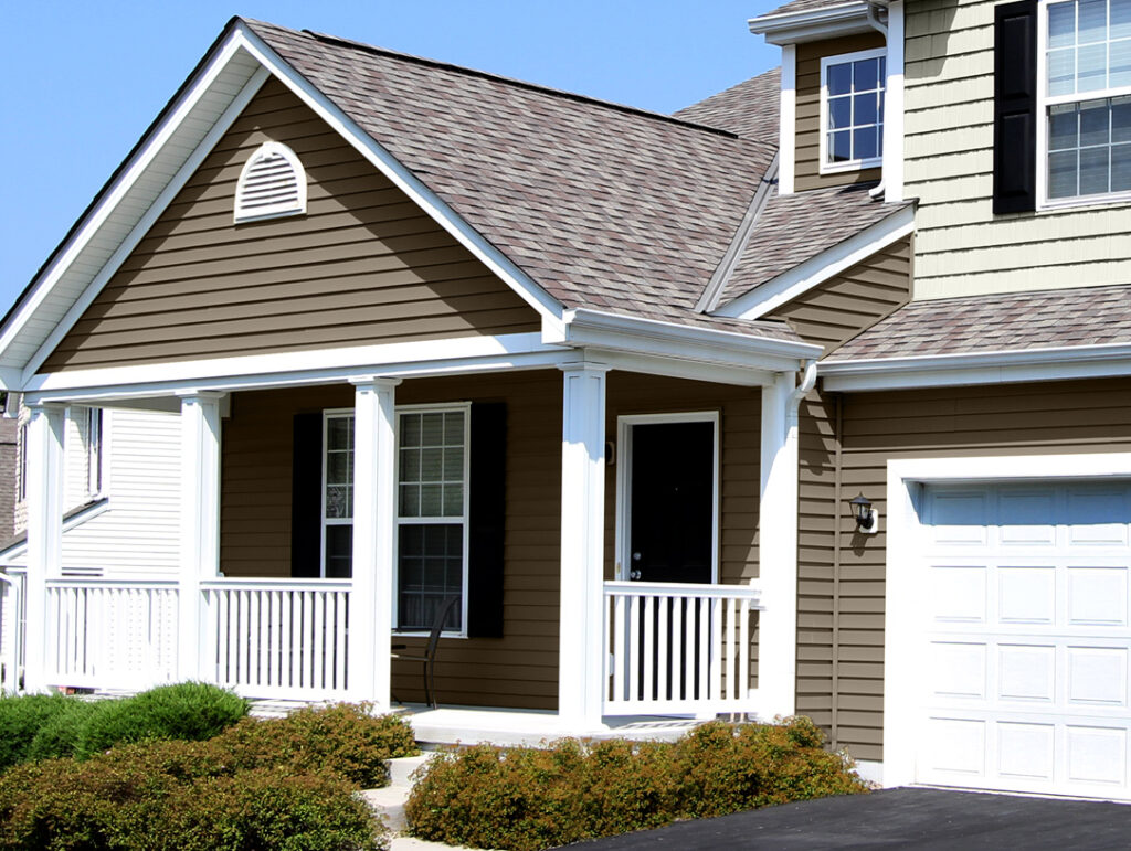 A suburban house with brown vinyl siding, white trim, a covered front porch with white railings, multiple windows, and part of a garage visible. The driveway and some bushes are in the foreground.