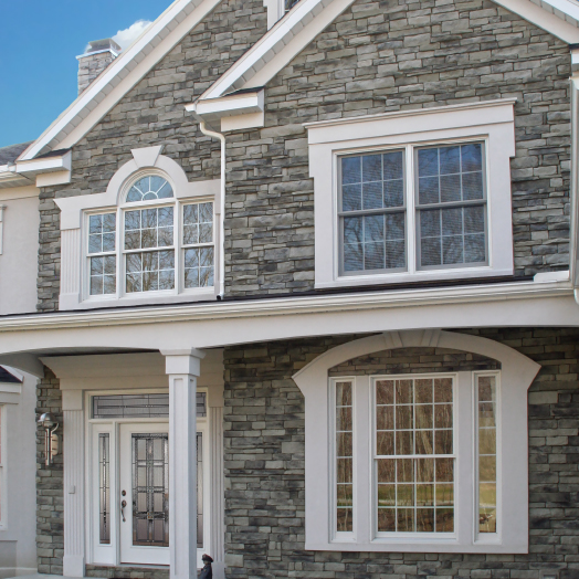 A close-up of a house exterior features gray stone siding, large white-trimmed windows, and a glass front door with decorative designs, under a white porch roof with columns.