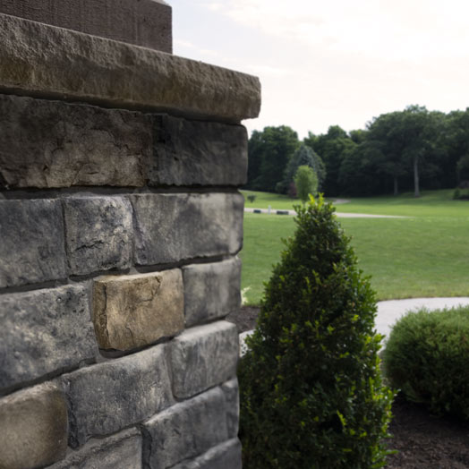 A close-up of a stone wall on the left side, with green bushes and an open park with grass and trees in the background under a partly cloudy sky.