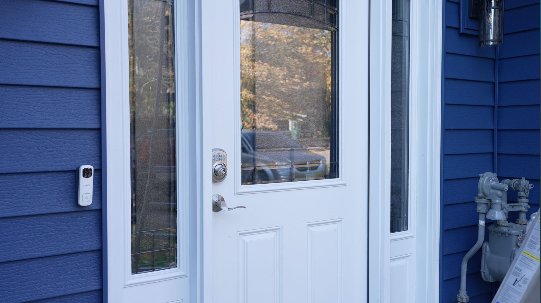 White entry door with glass panels on a blue house exterior. A silver door handle and smart lock are visible, along with a video doorbell and gas meters nearby. Reflections of trees appear in the glass. Perfect example of modern Entry Doors style.