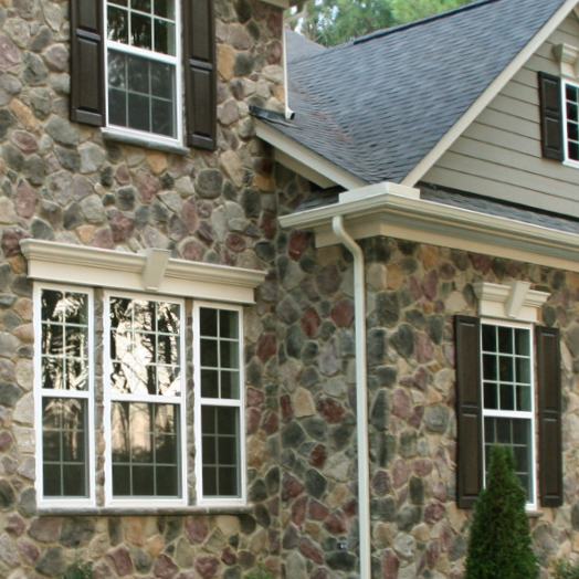 A stone house exterior with white-trimmed windows, dark brown shutters, and a gray shingled roof. A small evergreen shrub is in front of the building.