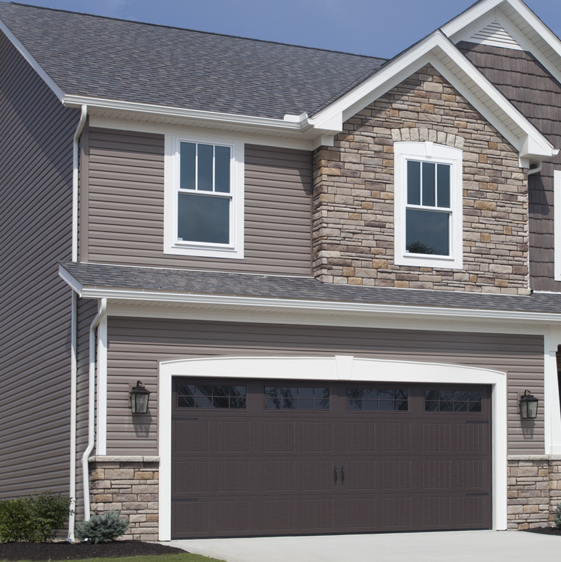 Two-story suburban house with light brown vinyl siding, stone accents, white trim, and a dark brown double garage door. Two lantern-style lights are mounted beside the garage. The concrete driveway leads up to the home.