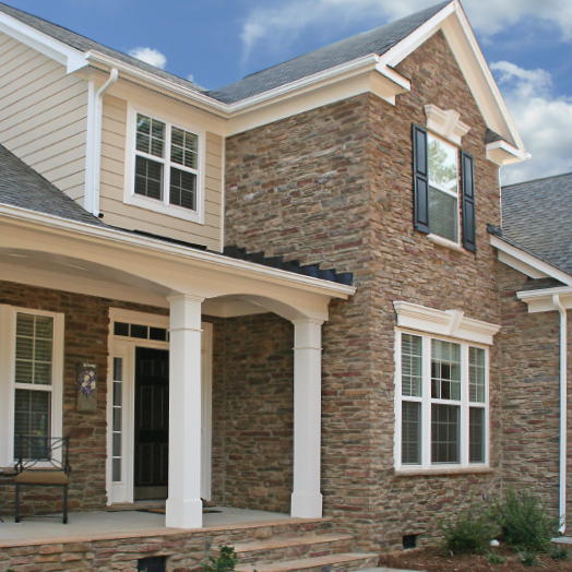 A modern house with a stone exterior, black shutters, white trim, a covered front porch with columns, and several large windows. The sky is blue with a few clouds.