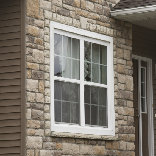 A close-up of a house exterior featuring a white-framed window set in a stone wall. Sheer curtains are visible through the window. Part of a door and the roof edge are also visible.