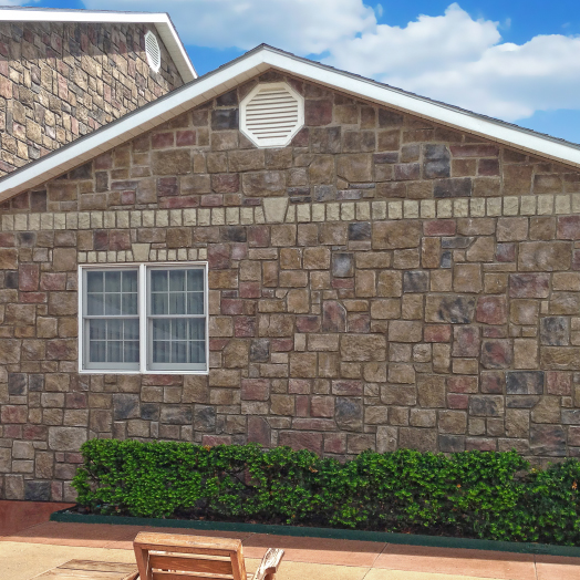 A house with a stone exterior wall, white-trimmed window, and a small vent near the roof. There are green bushes along the wall and a wooden chair in the foreground under a blue sky with clouds.