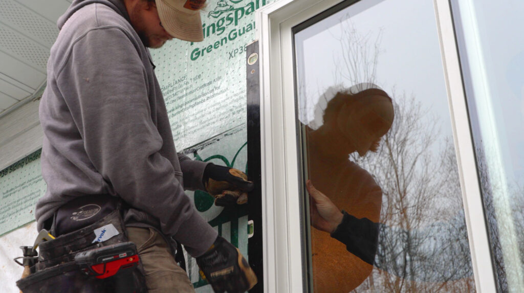 A worker in a cap and hoodie installs a window, possibly near future patio doors, on a house under construction. Using tools, the worker’s reflection is clearly visible in the glass of the window.