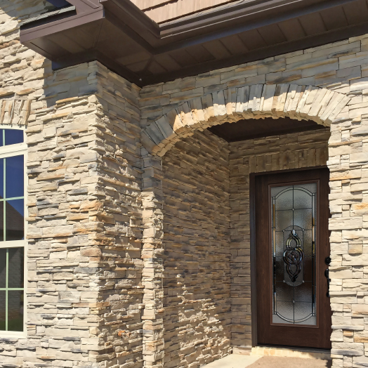 Stone exterior of a house with an arched doorway and a decorative glass front door. Sunlight casts shadows on the textured stone walls. A window is visible to the left of the door.