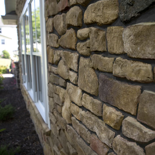Close-up view of a house exterior wall made of irregularly shaped tan and brown stones with visible mortar between them, next to white-framed windows and landscaping in the background.