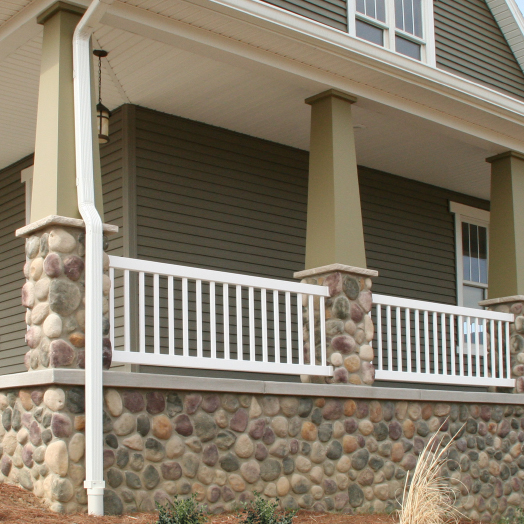 A house porch with beige pillars, a white railing, and a stone foundation featuring various colored stones. The house has olive-green siding and white trim. Grass and mulch are visible in the foreground.