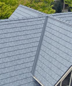A close-up view of a gray shingled roof with multiple peaks and valleys highlights its design, while lush green leafy trees form the background—showcasing an ideal setting for upgrading to durable metal roofing.