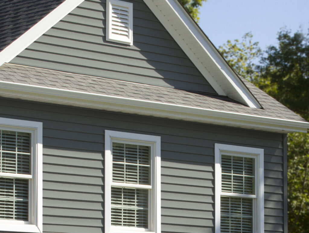 Close-up of a house exterior with gray vinyl siding, white trim, two windows with white frames and blinds, an attic vent, and part of a sloped roof visible, with trees and blue sky in the background.