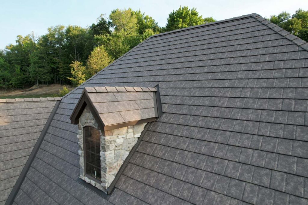 A close-up view of a house roof covered with dark gray shingles and featuring a small stone dormer with a window, highlighting the durability often associated with metal roofing. Green trees are visible in the background.