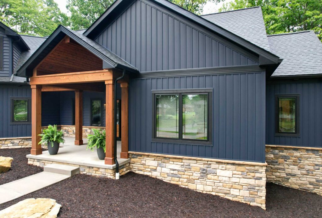 Modern house exterior renovation with dark blue vertical siding, stone accents, black-framed windows, and a wooden porch supported by columns. Two potted plants sit at the entrance, with mulch landscaping in the front.