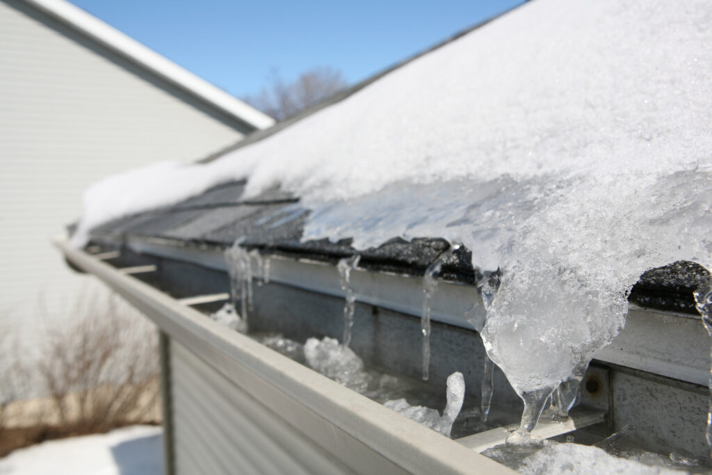 Ice and snow buildup on a house roof and gutter with icicles hanging down, seen on a clear, sunny winter day.