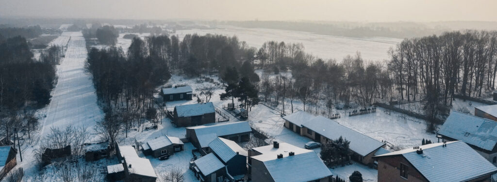 Aerial view of a small village with snow-covered rooftops and trees on a winter day, fields stretching into the distance under a pale, hazy sky with the sun faintly visible.