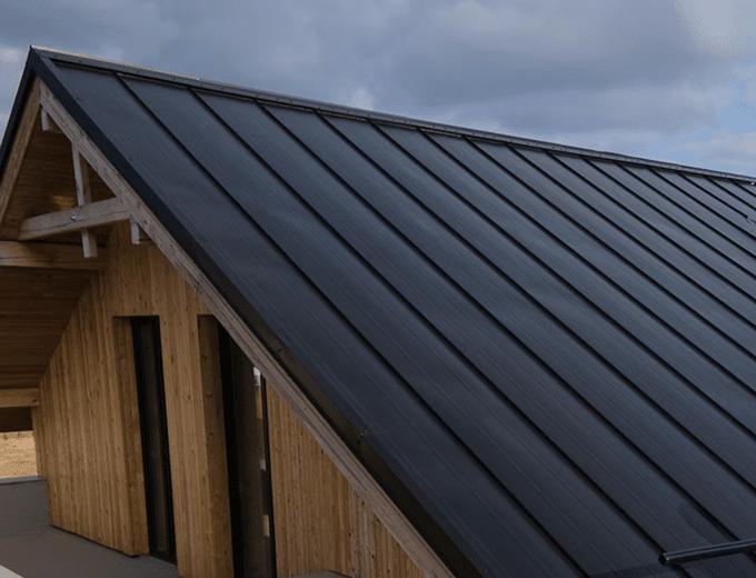 A close-up view of a slanted wooden house roof with integrated black solar panels under a cloudy sky.